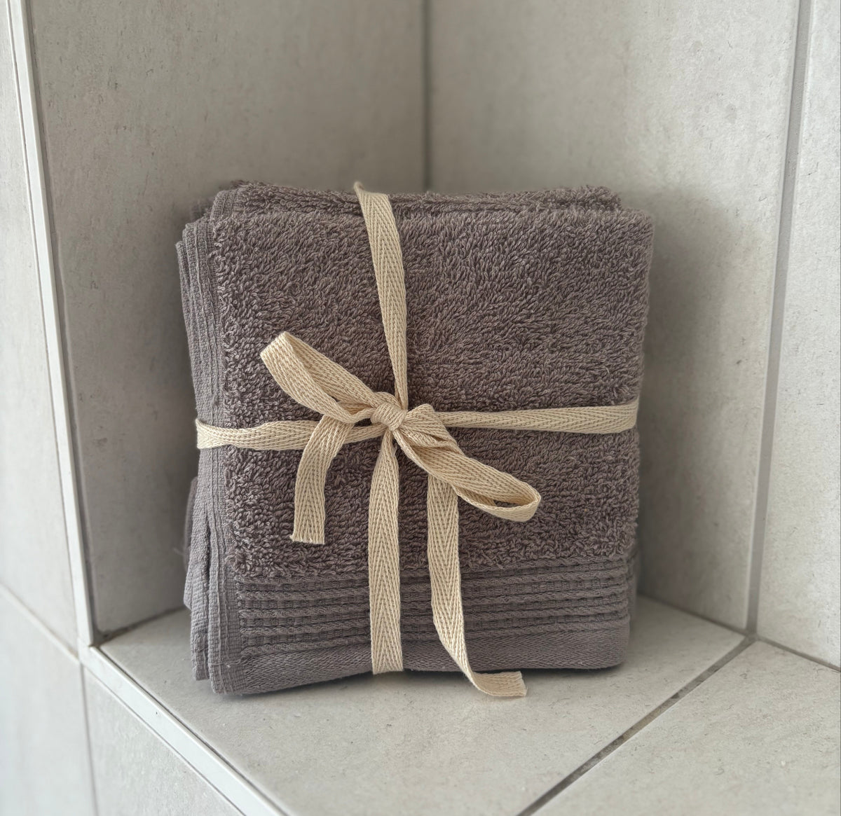 A stack of folded gray terry towels tied with a beige ribbon bow, placed on a light stone shelf in a tiled corner.
