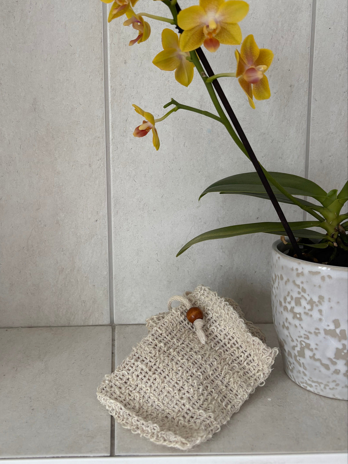 A small cream-colored woven pouch with a wooden bead closure rests on a light stone shelf next to a potted yellow orchid in a white speckled pot.