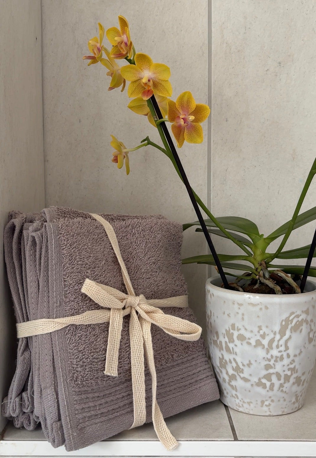 A stack of folded gray towels tied with a cream ribbon sits on a tiled shelf next to a potted yellow orchid.