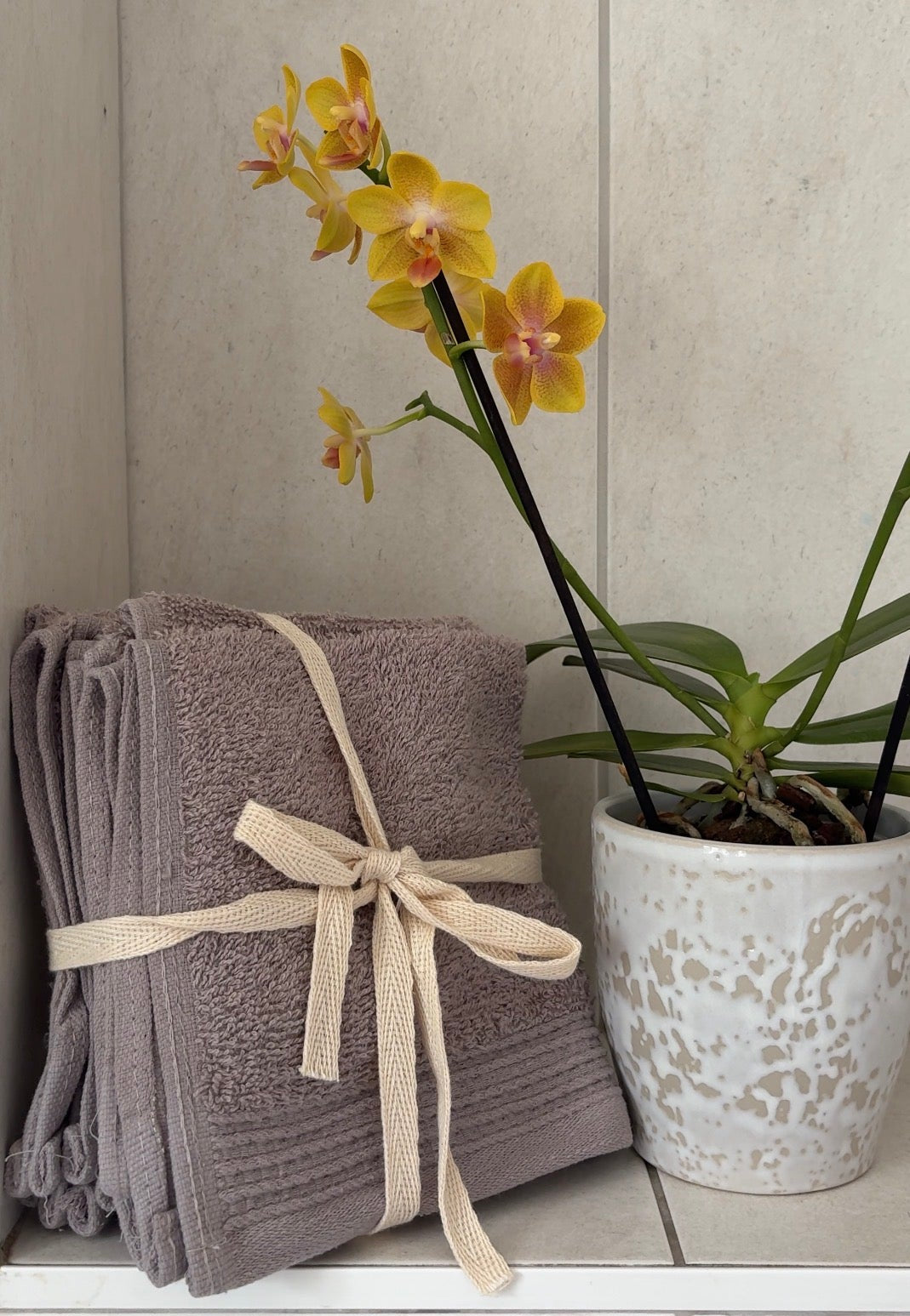 A stack of folded gray towels tied with a cream ribbon sits on a tiled shelf next to a potted yellow orchid.