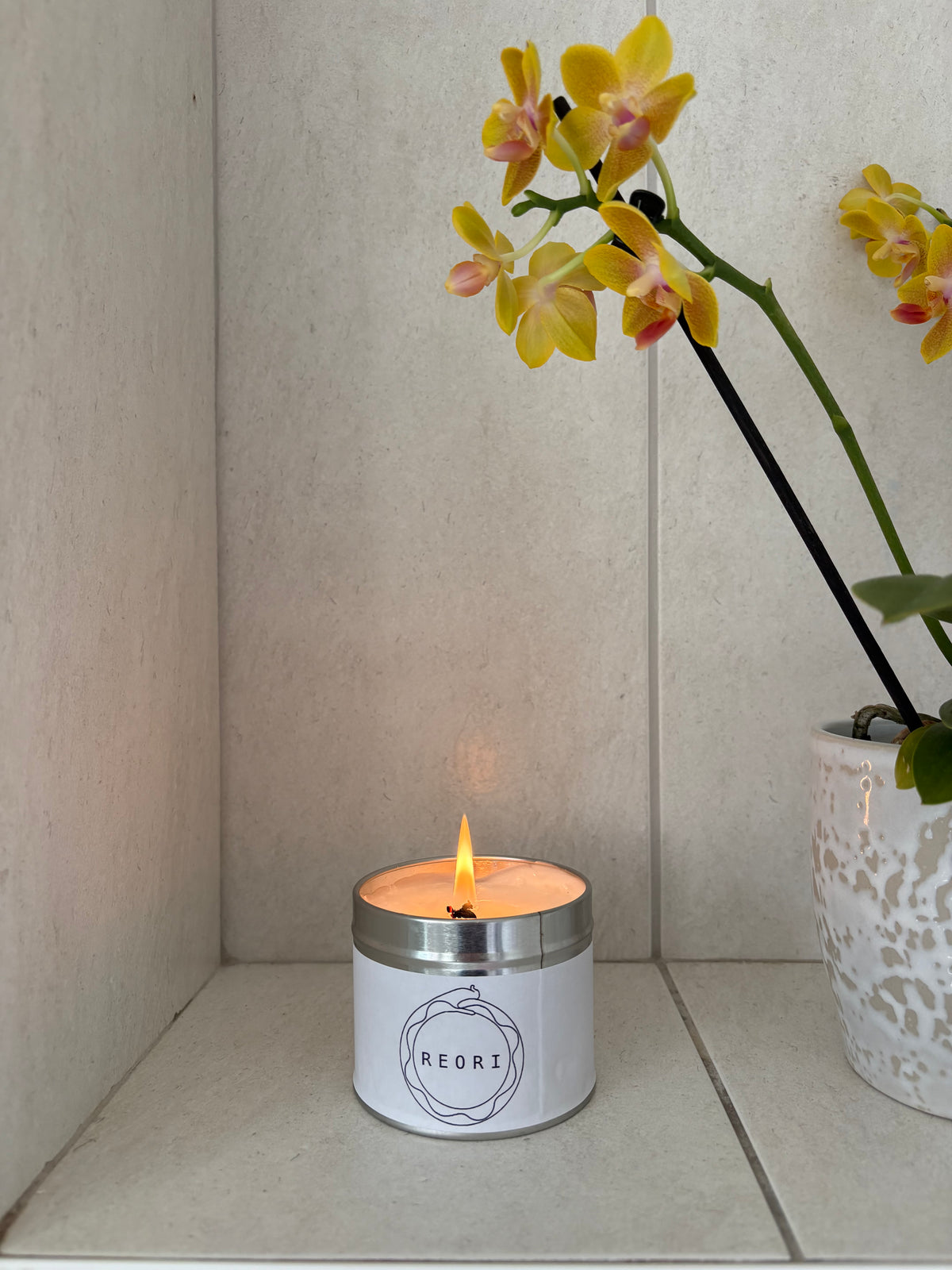 A white jar candle with a silver metal rim and the brand name 'REORI' printed on the front, lit with a warm flame. It sits on a beige tiled shelf next to a white ceramic pot holding yellow orchids.