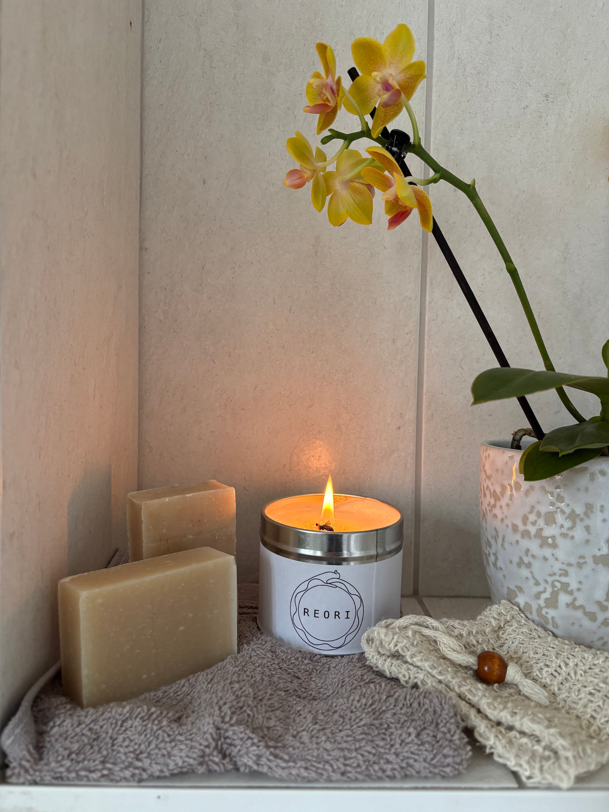 A lit white candle with a silver rim and the brand name 'REORI' on the front, styled on a shelf with two beige soap bars, a grey towel, a cream soap pouch, and a potted yellow orchid.
