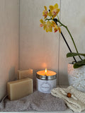 A lit white candle with a silver rim and the brand name 'REORI' on the front, styled on a shelf with two beige soap bars, a grey towel, a cream knit cloth, and a potted yellow orchid.