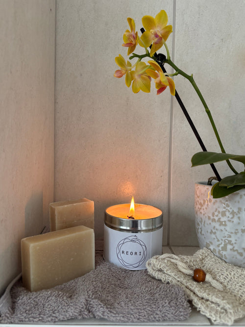 A lit white candle with a silver rim and the brand name 'REORI' on the front, styled on a shelf with two beige soap bars, a grey towel, a cream knit cloth, and a potted yellow orchid.