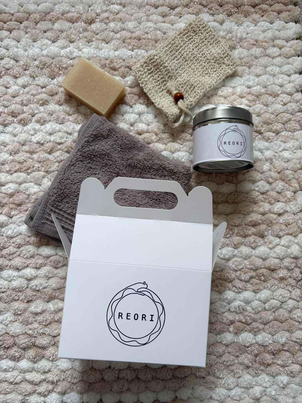 A curated bath and body gift set from REORI, including a white gift box, a gray towel, a beige soap bar, a beige scrubber, and a silver tin candle, all arranged on a textured cream and beige knit blanket.