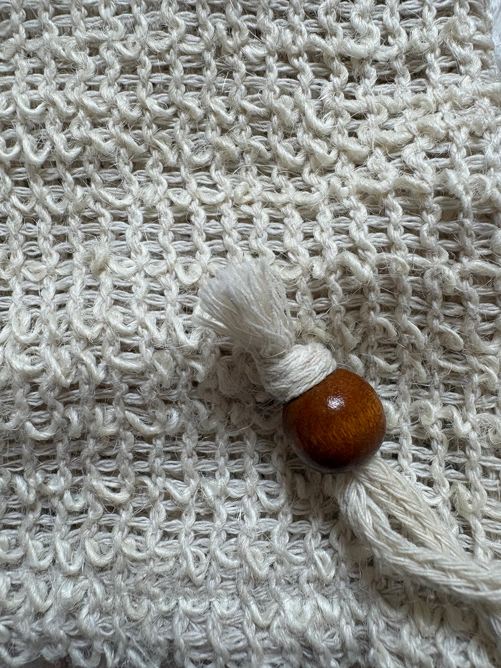 A close-up of a cream-colored textured soap pouch featuring a small decorative tassel charm with a polished brown wooden bead.