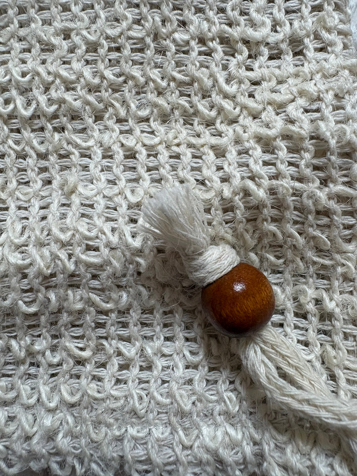 A close-up of a cream-colored textured soap pouch featuring a small decorative tassel charm with a polished brown wooden bead.