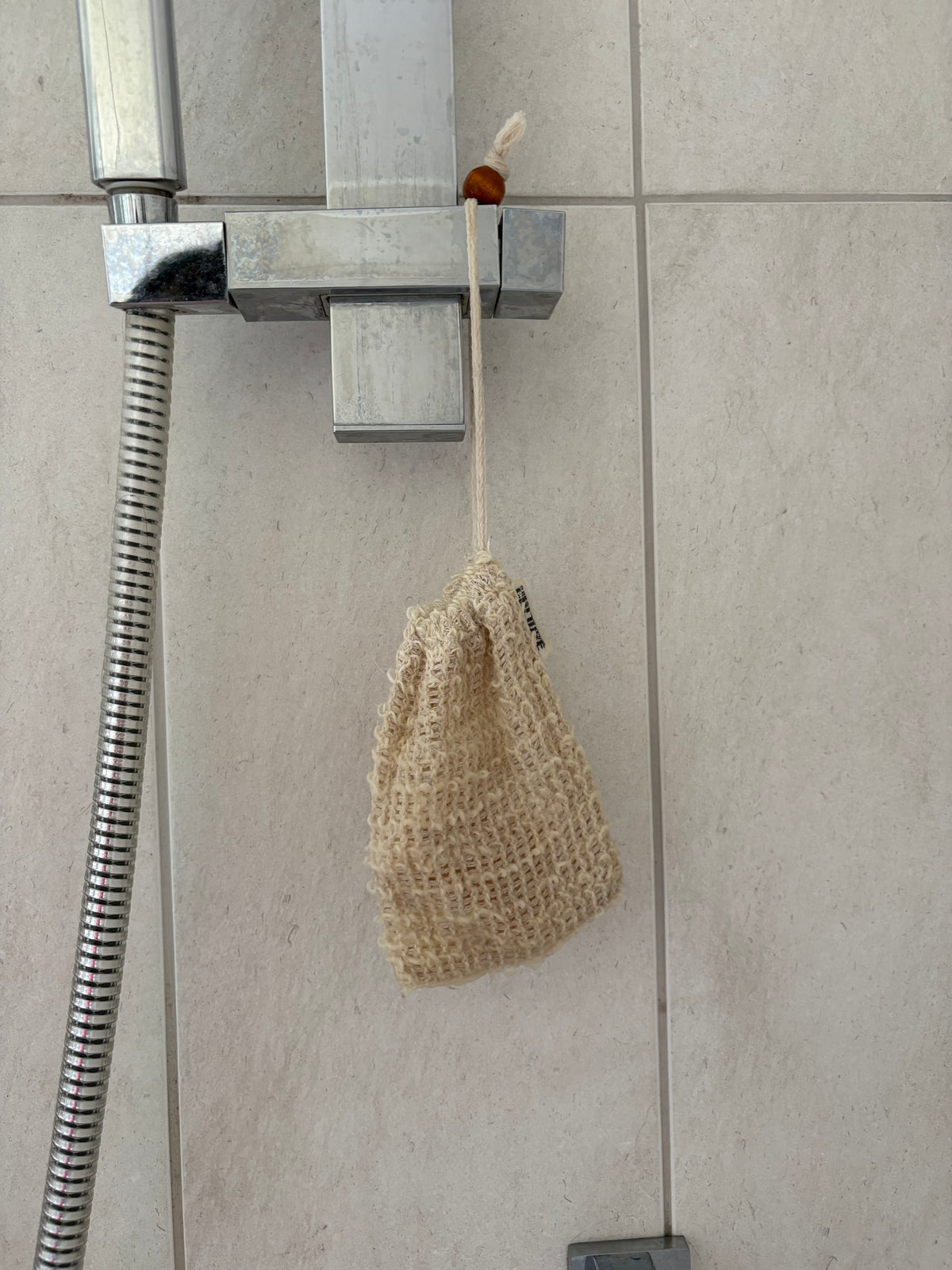 A beige woven drawstring bath sponge hanging from a chrome shower valve on a tiled bathroom wall.