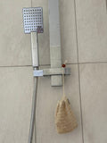 A wall-mounted shower fixture with a rectangular chrome shower head, flexible hose, and wall bracket installed on beige tiled bathroom walls. A beige mesh loofah hangs from the hose.