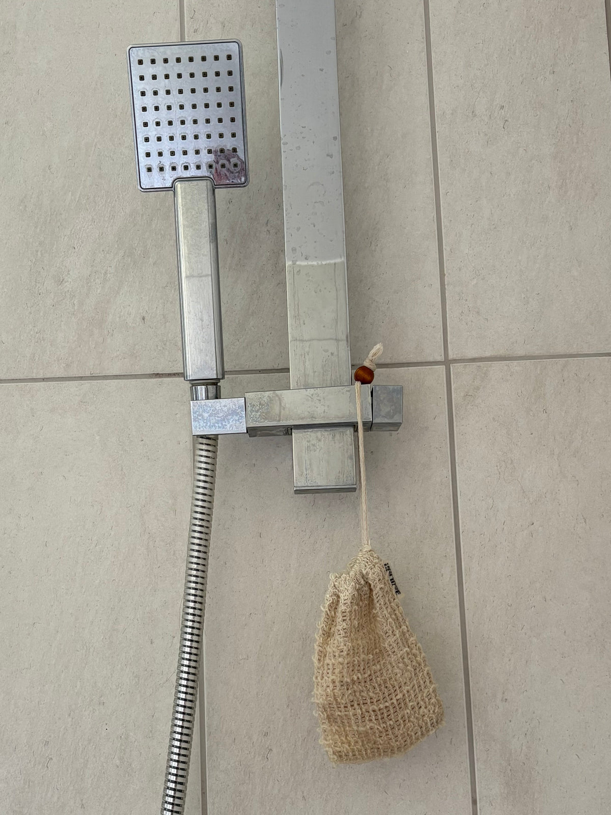 A wall-mounted shower fixture with a rectangular chrome shower head, flexible hose, and wall bracket installed on beige tiled bathroom walls. A beige mesh loofah hangs from the hose.
