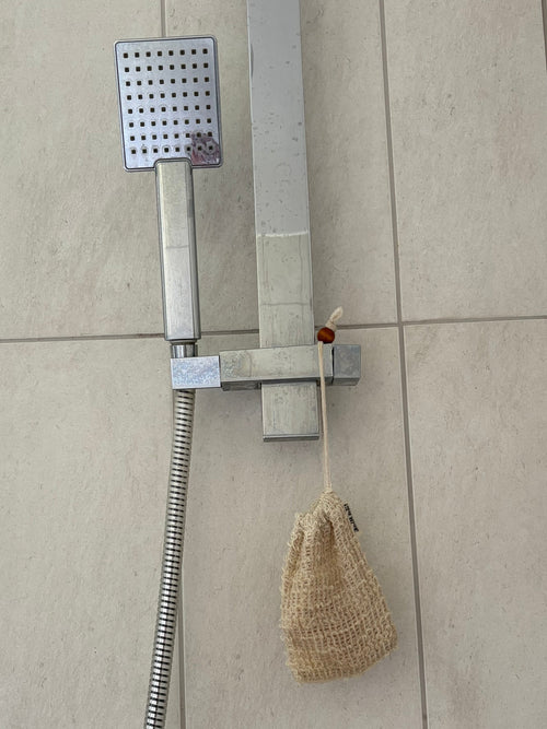 A wall-mounted shower fixture with a rectangular chrome shower head, flexible hose, and wall bracket installed on beige tiled bathroom walls. A beige mesh loofah hangs from the hose.