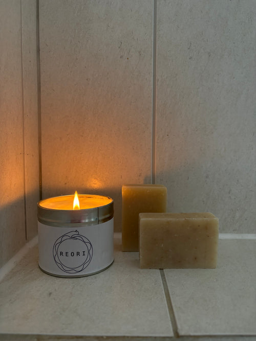 A lit white candle in a silver tin labeled REORI sits on a tiled surface next to two rectangular beige soap bars against a light stone wall.