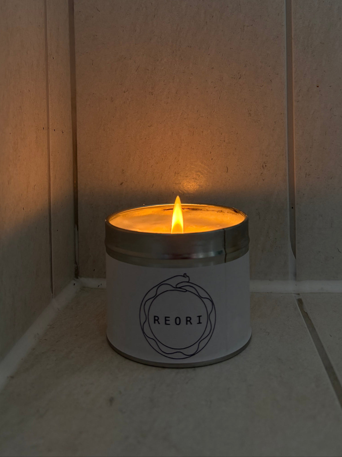 A lit REORI candle in a white tin with a silver metal rim, sitting on a stone floor against a stone wall.