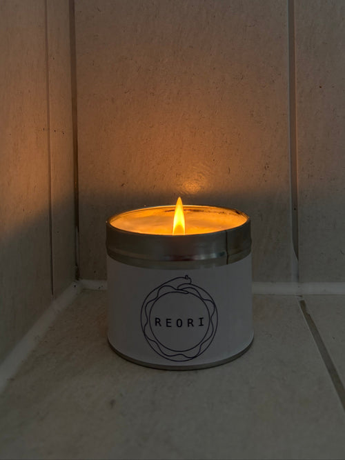 A lit REORI candle in a white tin with a silver metal rim, sitting on a stone floor against a stone wall.