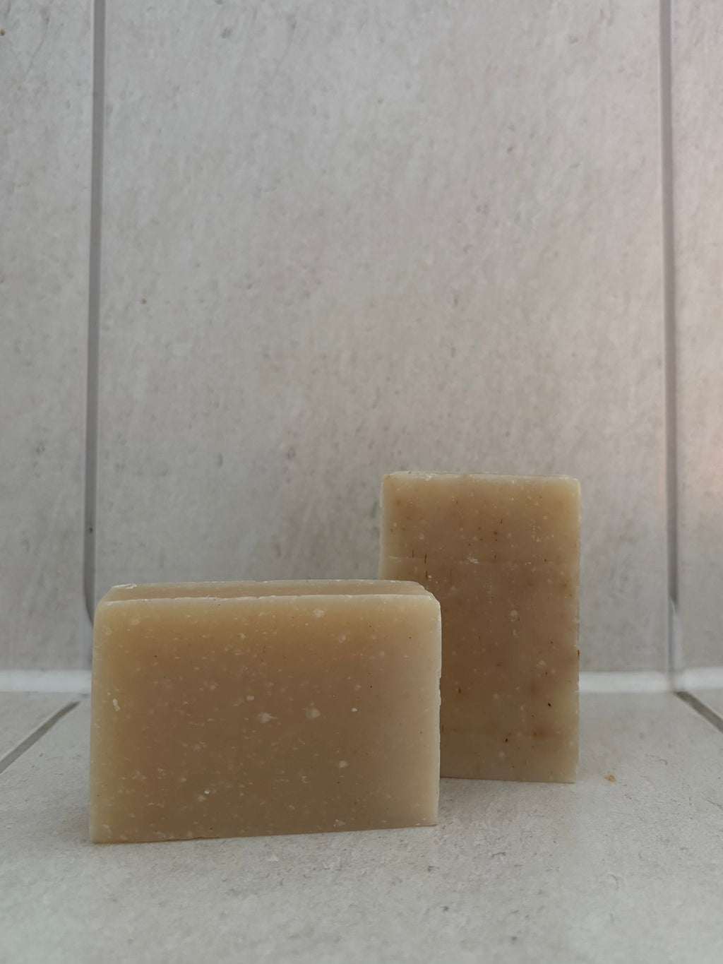 Two rectangular bars of natural beige soap standing upright on a light tiled surface against a matching tiled background.