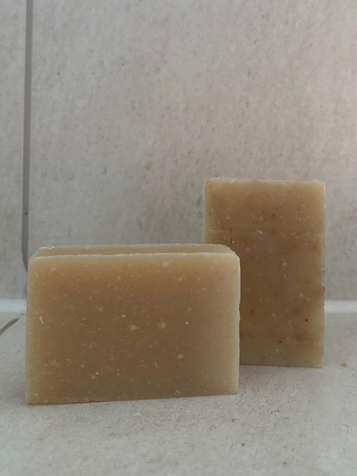 Two rectangular bars of natural beige soap standing upright on a light tiled surface against a matching tiled background.