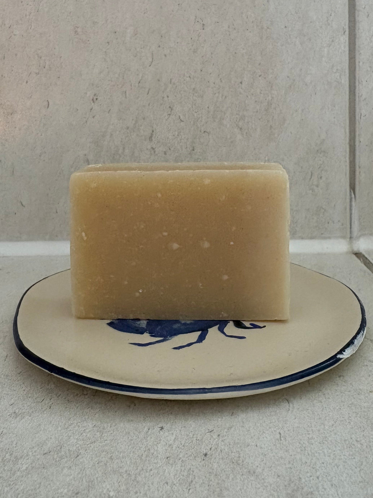 A rectangular bar of pale beige soap resting on a small round ceramic dish with a blue floral design, set against a light gray tiled background.