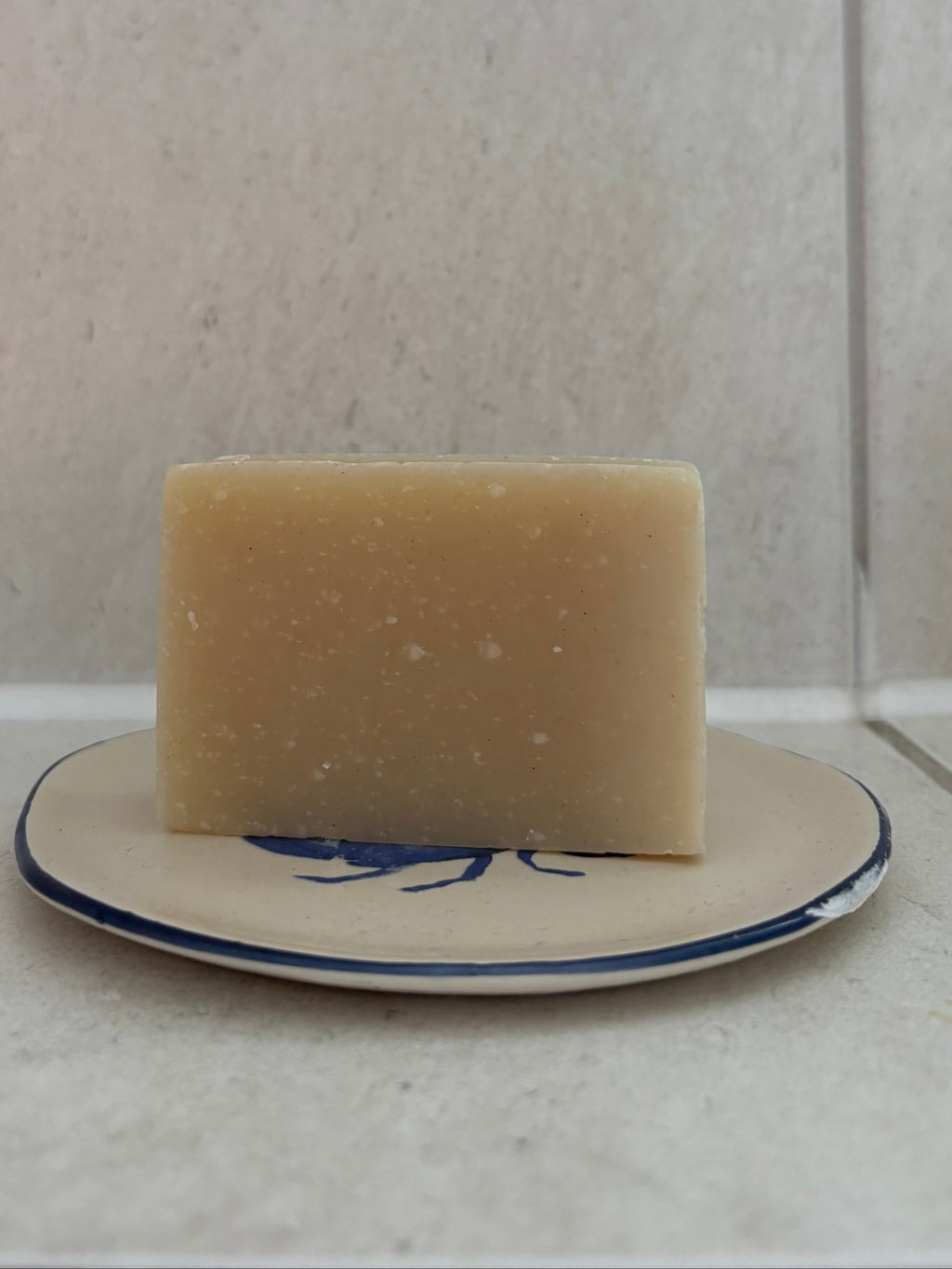 A rectangular bar of pale beige soap standing upright on a small white dish with a blue floral border, set against a light tiled background.