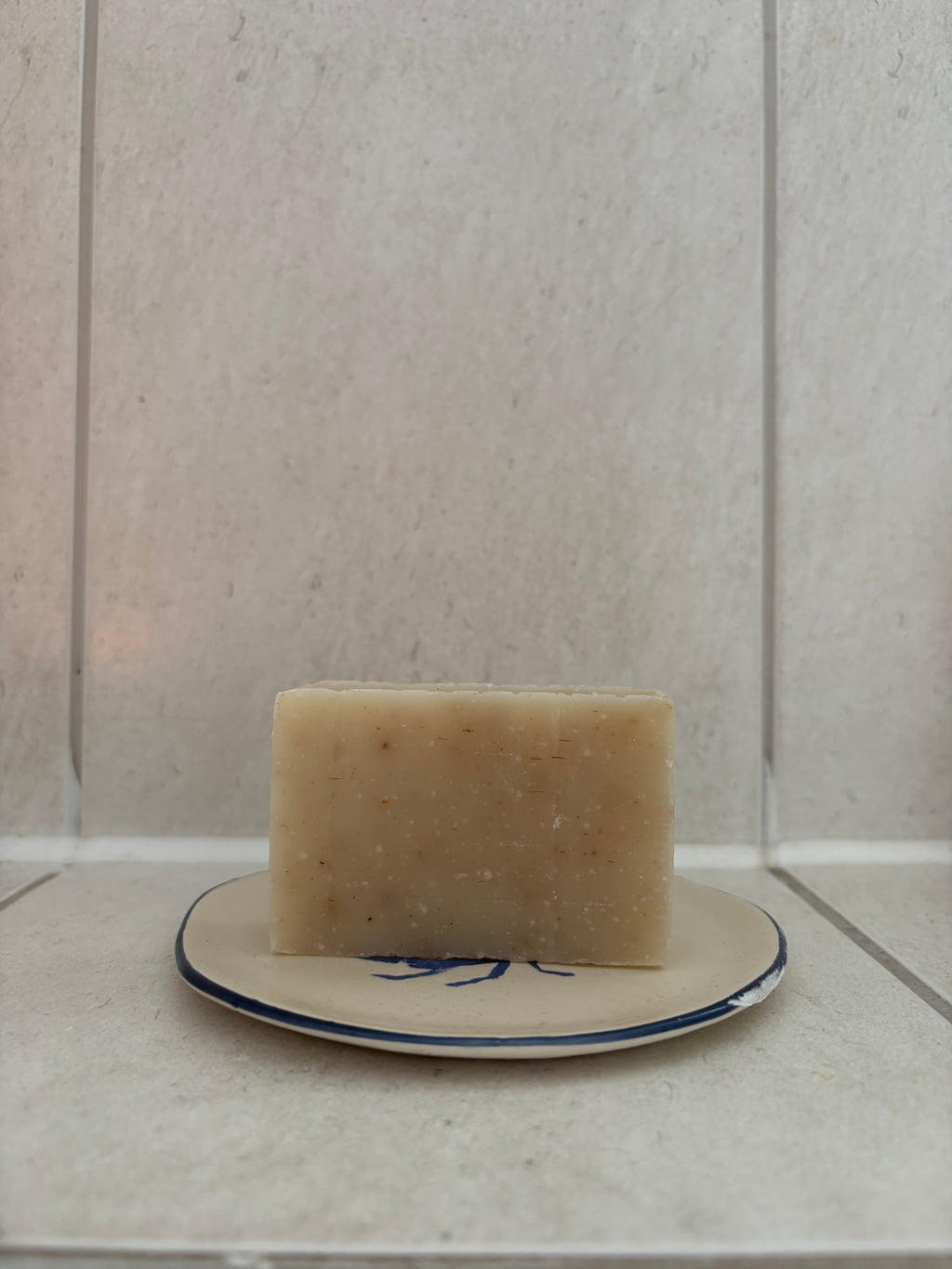 A rectangular bar of natural beige soap with visible botanical inclusions, resting on a small beige ceramic dish with a blue rim against a light beige tiled background.