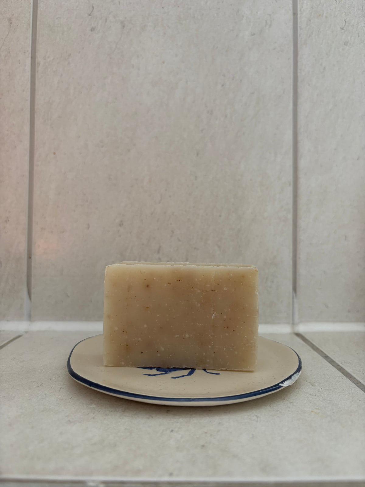 A rectangular bar of natural beige soap with visible botanical inclusions, resting on a small beige ceramic dish with a blue rim against a light beige tiled background.