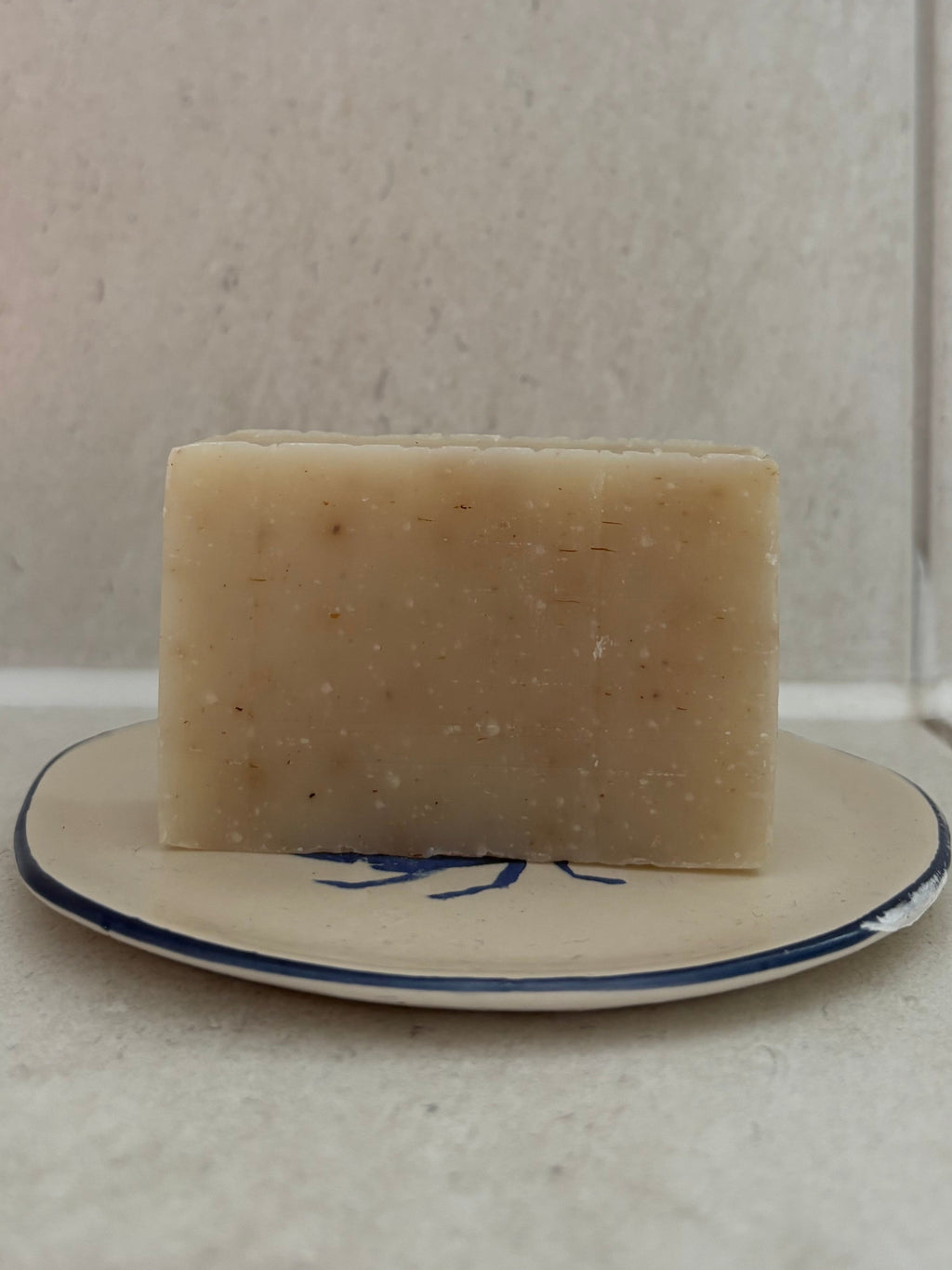 A rectangular bar of pale beige soap with visible specks, resting on a small white plate with a blue rim against a light tiled background.