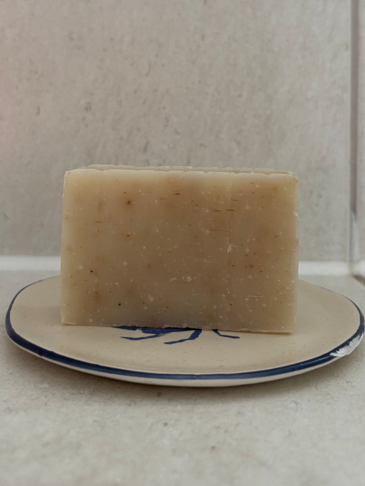 A rectangular bar of pale beige soap with visible specks, resting on a small white plate with a blue rim against a light tiled background.