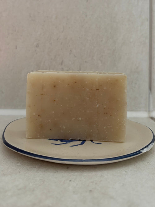 A rectangular bar of pale beige soap with visible specks, resting on a small white plate with a blue rim against a light tiled background.