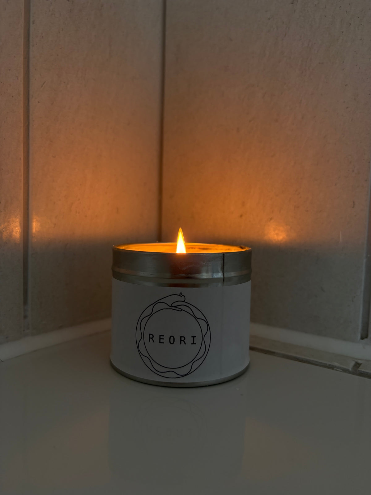 A lit REORI scented candle in a white metal tin with a silver lid, sitting on a white surface in the corner of a room with warm ambient lighting.
