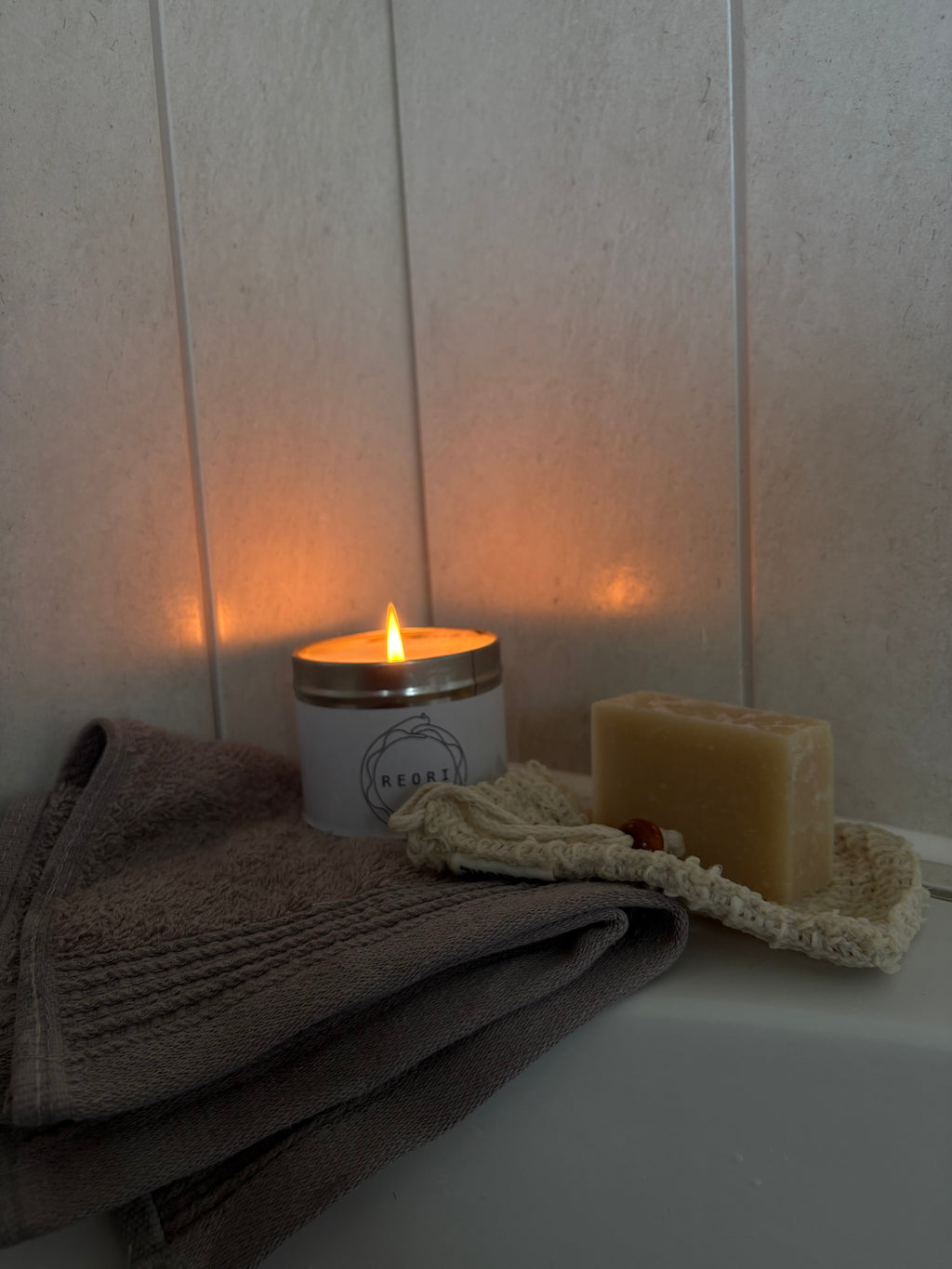 A lit white candle with a gold-toned lid sits on a white surface next to a folded grey towel and a bar of soap on a textured cream cloth, against a light beige panelled wall.
