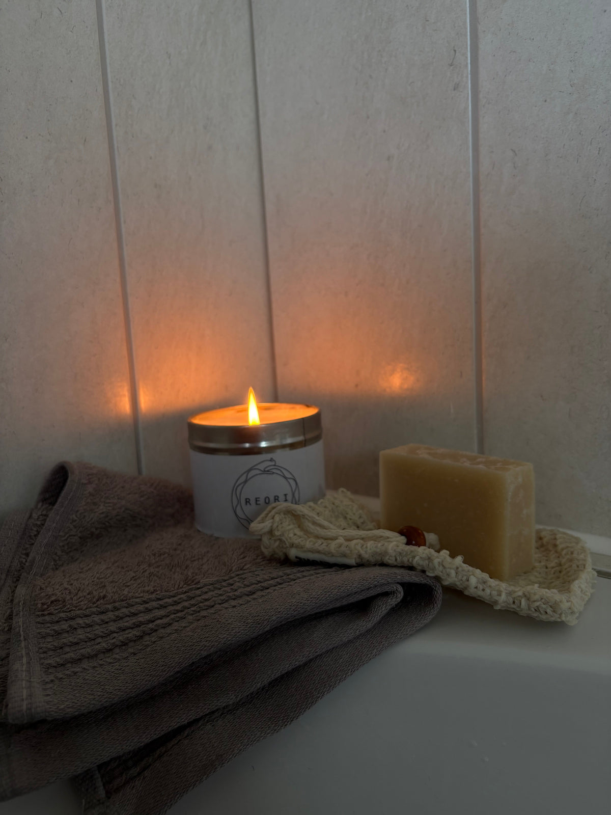 A lit white candle with a gold-toned lid sits on a white surface next to a folded grey towel and a bar of soap on a textured cream cloth, against a light beige panelled wall.