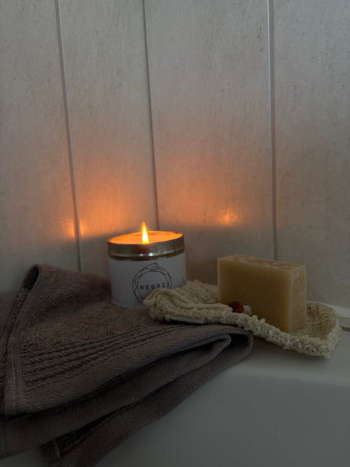 A lit white candle with a gold-toned lid sits on a white surface next to a folded grey towel and a bar of soap on a textured cream cloth, against a light beige panelled wall.